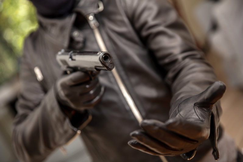 Armed robber with a pistol in gloved hand. Gunman wearing black leather jacket aiming with a gun, asking for money, closeup view.